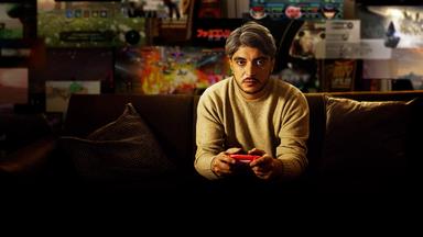 Host Khesrau Behroz sitzt mit Videospiel-Controller auf dem Sofa.