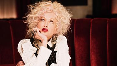 Cyndi Lauper sitzt auf einem roten Sessel, den Kopf auf der Hand abgestützt. Sie schaut nach oben.