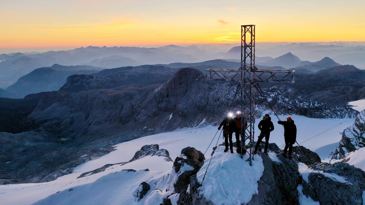 Bergsteiger erreichen bei Sonnenaufgang das Gipfelkreuz des Dachsteins, umgeben von einer atemberaubenden Hochgebirgslandschaft.