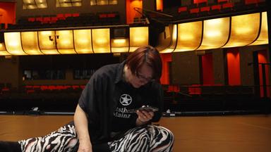 Tänzer schaut sich vor der Probe nochmal die Choreografie an. Er sitzt auf einer dunklen Bühne , während er mit dem Handy in der Hand und Paieren auf dem Boden sich die Schritte einprägt.