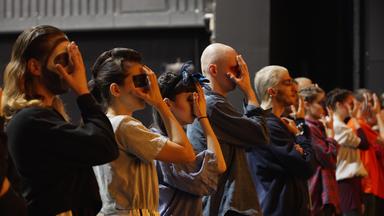 Eine Gruppe von Tänzer*innen steht gestaffelt in einer Reihe auf einer Bühne. Sie tragen Alltags- und Trainingskleidung. Ihre Gesichter sind geschminkt, manche bunt, manche schwarz-weiß. Alle halten ihre rechte Hand vors Gesicht.  