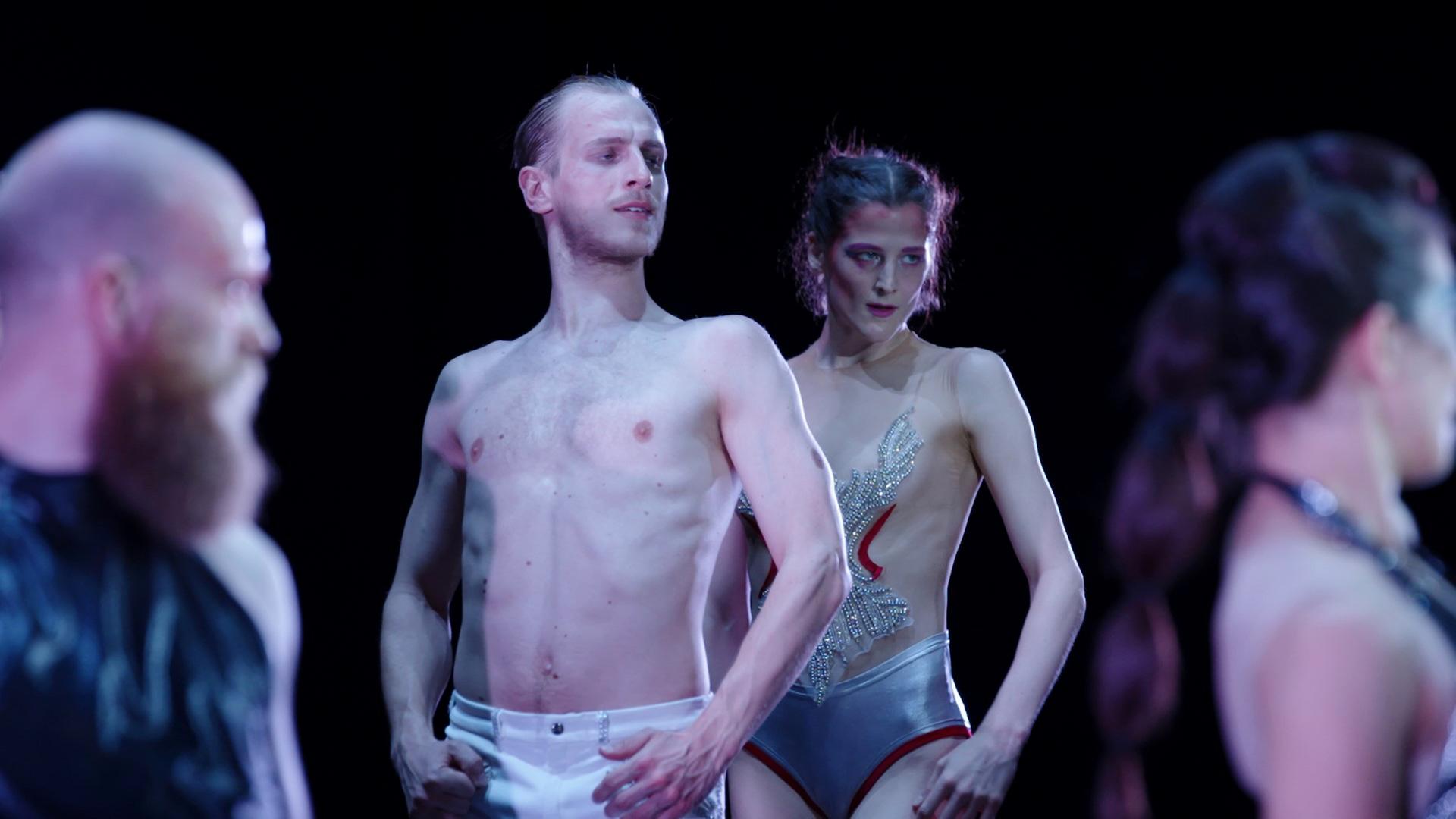 Auf einer dunklen Bühne stehen vier Personen, zwei Männer und zwei Frauen. Der Mann am rechten und die Frau am linken Bildrand sind nur unscharf zu erkennen. In der Mitte steht links ein junger Mann mit nacktem Oberkörper, er trägt eine weiße Hose, versetzt leicht dahinter, rechts von ihm steht eine junge Frau in einem Trikot mit silberner Hose und einem transparenten Oberteil, das mit glitzernden Steinen bestickt ist. Sie stehen aufrecht und blicken nach rechts mit strengem Blick aus dem Bild.