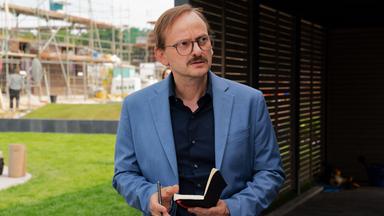 Vor dem Haus der Bressins. Danowski (Milan Peschel) hält sein Notizbuch in der Hand und blickt misstrauisch drein.