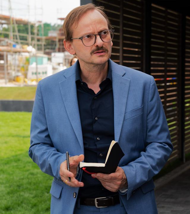 Vor dem Haus der Bressins. Danowski (Milan Peschel) hält sein Notizbuch in der Hand und blickt misstrauisch drein.