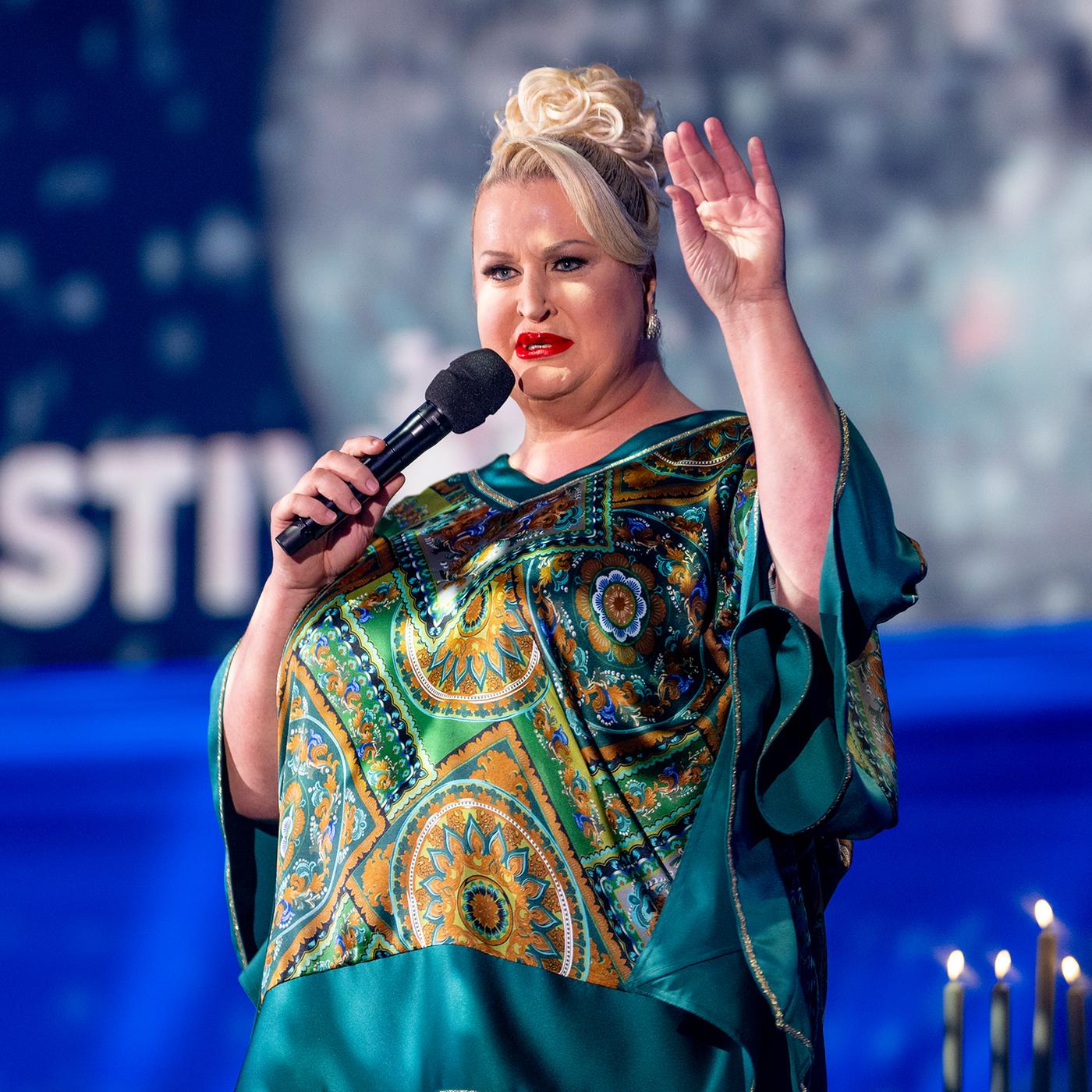 Daphne Deluxe steht in der Mitte des Bildes. Sie trägt ein blaugrünes Oberteil und hebt ihre linke Hand. Der Hintergrund ist blau beleuchtet.