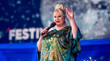 Daphne Deluxe steht in der Mitte des Bildes. Sie trägt ein blaugrünes Oberteil und hebt ihre linke Hand. Der Hintergrund ist blau beleuchtet.