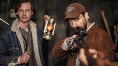 Zwei Männer in der Nacht; einer hält eine Laterne, der andere zielt mit einem Gewehr. 