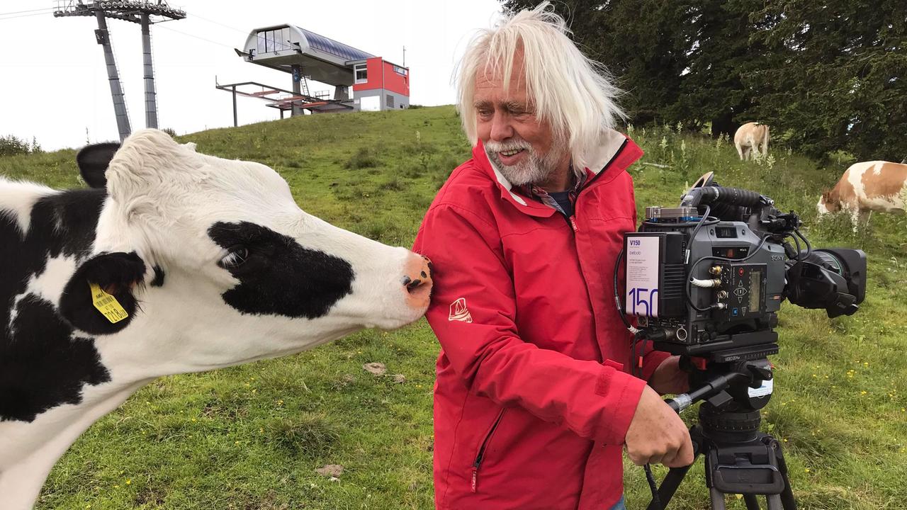 Im Bild ist ein Naturfilmer mit grauen Haaren und einem roten Mantel zu sehen. Er steht neben einer schwarz-weißen Kuh und hält eine Kamera. Im Hintergrund sind Wiesen und Kühe.