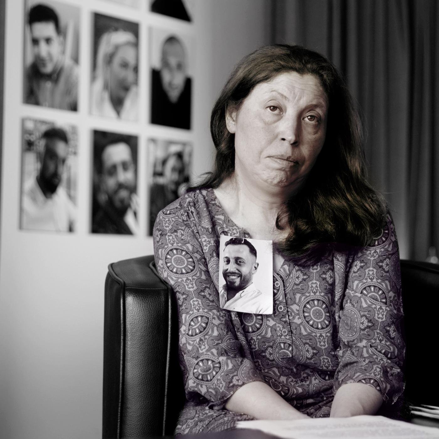 Frau blickt direkt in die Kamera. An ihrem Revers ist das Foto eines jungen Mannes befestigt.