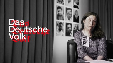 Frau blickt direkt in die Kamera. An ihrem Revers ist das Foto eines jungen Mannes befestigt.