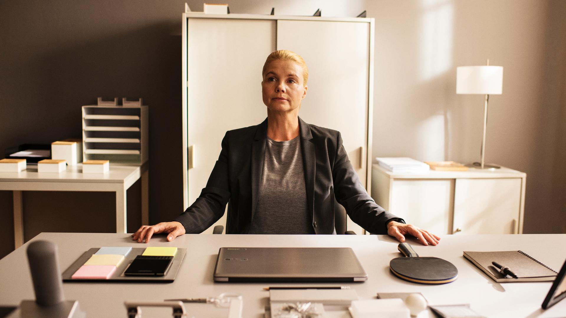 "Ella Schön - Das Ding mit der Liebe":Ella (Annette Frier) sitzt an einem sehr ordentlichen Schreibtisch in einem Büro und hat beide Hände auf den Tisch gelegt. Vor ihr auf dem Tisch befinden sich Schreibwaren, ein Laptop, ein Handy und ein Tischtennisschläger, alles wohlgeordnet.