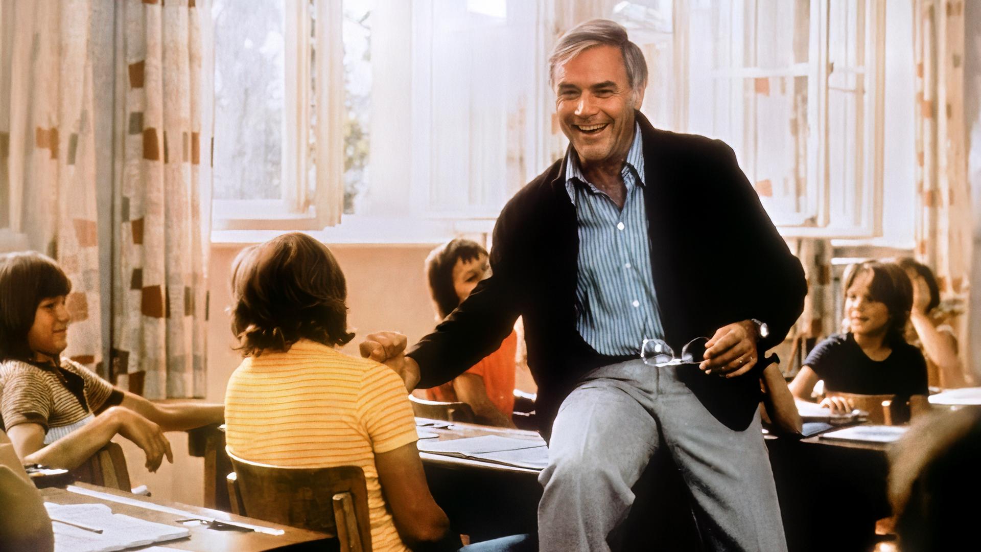 Dr. Johannes Bökh (Joachim Fuchsberger) sitzt auf einem Tisch im Klassenzimmer und lacht seine Schüler an.
