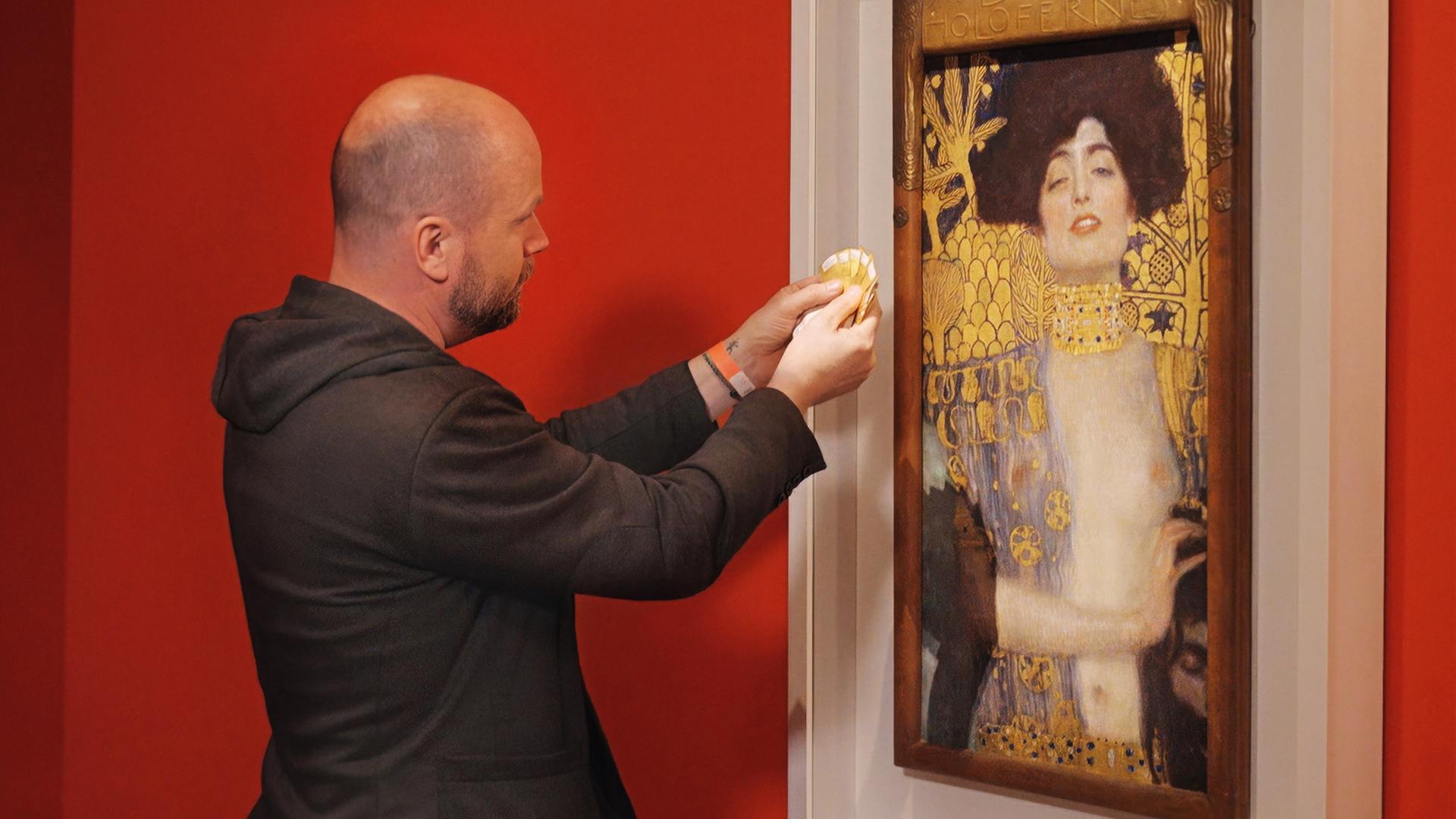 Ein Mann hält Farbmusterkarten an das Gemälde Judith und Holofernes von Gustav Klimt, das in einem weißen Rahmen vor einer roten Wand hängt.