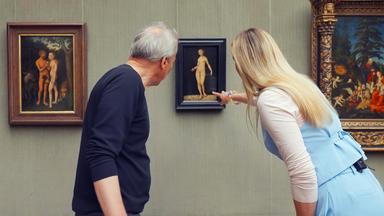Ein älterer Mann und eine Frau betrachten ein Gemälde von Lucas Cranach, das Adam und Eva zeigt, in einer Gemäldegalerie.