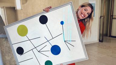 Frau in rotem Anzug posiert lachend mit einem großformatigen abstrakten Gemälde in einem hellen Museumsflur.