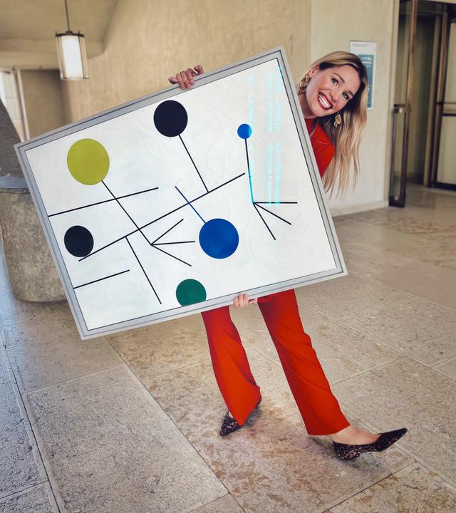 Frau in rotem Anzug posiert lachend mit einem großformatigen abstrakten Gemälde in einem hellen Museumsflur.