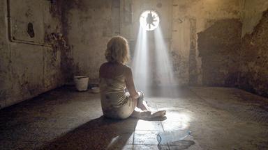 Eine Frau in einem beschmutzten Unterkleid sitzt am Boden eines schmutzigen Kellerraums. Durch einen Lüftungsschacht fällt Sonnenlicht. Im Vordergrund liegt eine leere Kunststoffflasche auf dem Boden. Im Hintergrund stehen ein Eimer und eine Schüssel auf dem Boden.