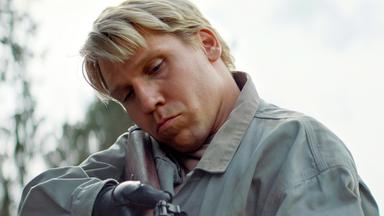 Ein blonder Mann mit einem Gewehr im Anschlag zielt auf etwas.