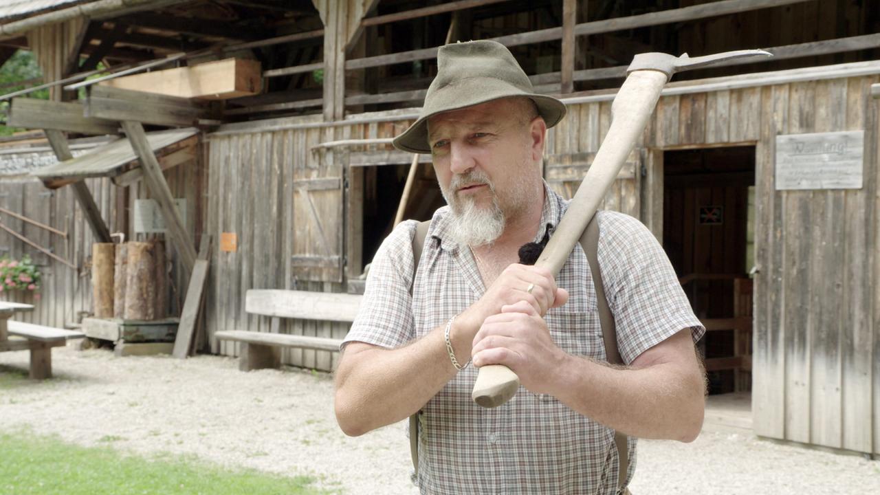 Ein älterer Mann mit grauem Bart steht vor einem traditionellen Holzhaus und hält eine Hacke in den Händen. Er trägt ein kariertes Hemd, Hosenträger und einen grünen Filzhut. Die Szene vermittelt eine ländliche, handwerklich geprägte Atmosphäre.