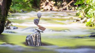 Mehrere kunstvoll aufeinandergetürmte Steine stehen am Ufer eines Flusses im Glantal, dahinter Wasser und grüne Vegetation.