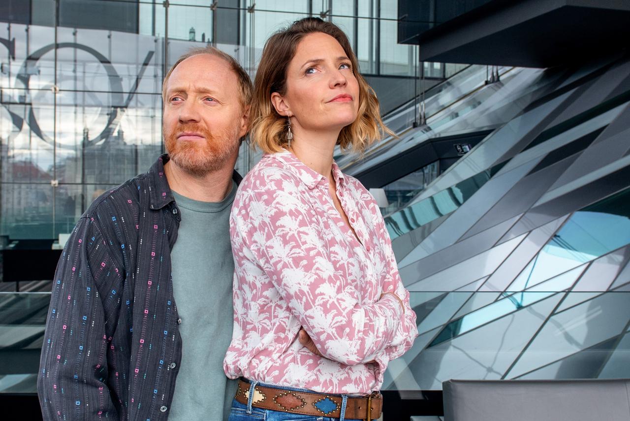 Das Bild zeigt zwei Personen, die eng beieinander stehen. Die erste Person, links im Bild, hat rotes Haar und trägt ein gestreiftes, dunkelblaues Hemd mit bunten Punkten. Die zweite Person, rechts, hat braune, lockige Haare und trägt eine helle Bluse mit einem Palmenmuster. Sie hat die Arme vor der Brust verschränkt und schaut leicht nach oben. Im Hintergrund ist eine moderne Architektur zu sehen, die durch große Glasflächen und geschwungene Linien charakterisiert ist. Das Licht fällt durch die Fenster und schafft eine helle, luftige Atmosphäre.