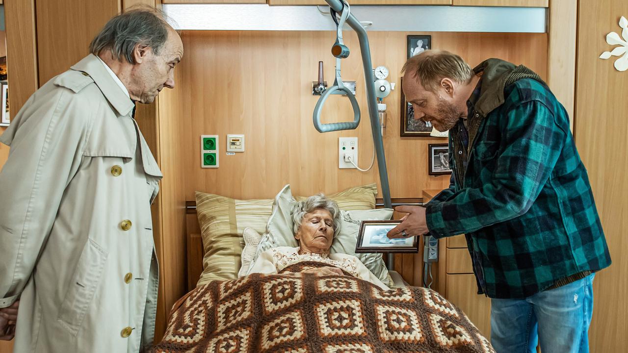 Das Bild zeigt eine Szene aus einem Krankenhauszimmer. In der Mitte liegt eine ältere Frau, die auf einem Bett mit einer karierten Decke liegt. Ihr Gesicht ist entspannt, und sie hat silbergraue Haare. Neben dem Bett ist ein Stützgriff installiert, und im Hintergrund sind einige Fotografien an der Wand zu sehen.  Links steht ein älterer Mann in einem beigen Trenchcoat, der einen nachdenklichen oder besorgten Gesichtsausdruck hat. Rechts kniet ein weiterer Mann in einer karierten Jacke und einer gestreiften Bluse, der der älteren Frau ein Bild zeigt. Das Zimmer hat Holzverkleidungen und eine freundliche, aber traurige Atmosphäre.   Die Szenerie vermittelt ein Gefühl von Dramatik und Emotion, da es um die Beziehung zwischen den Charakteren und eine nicht erfüllte Liebe geht.