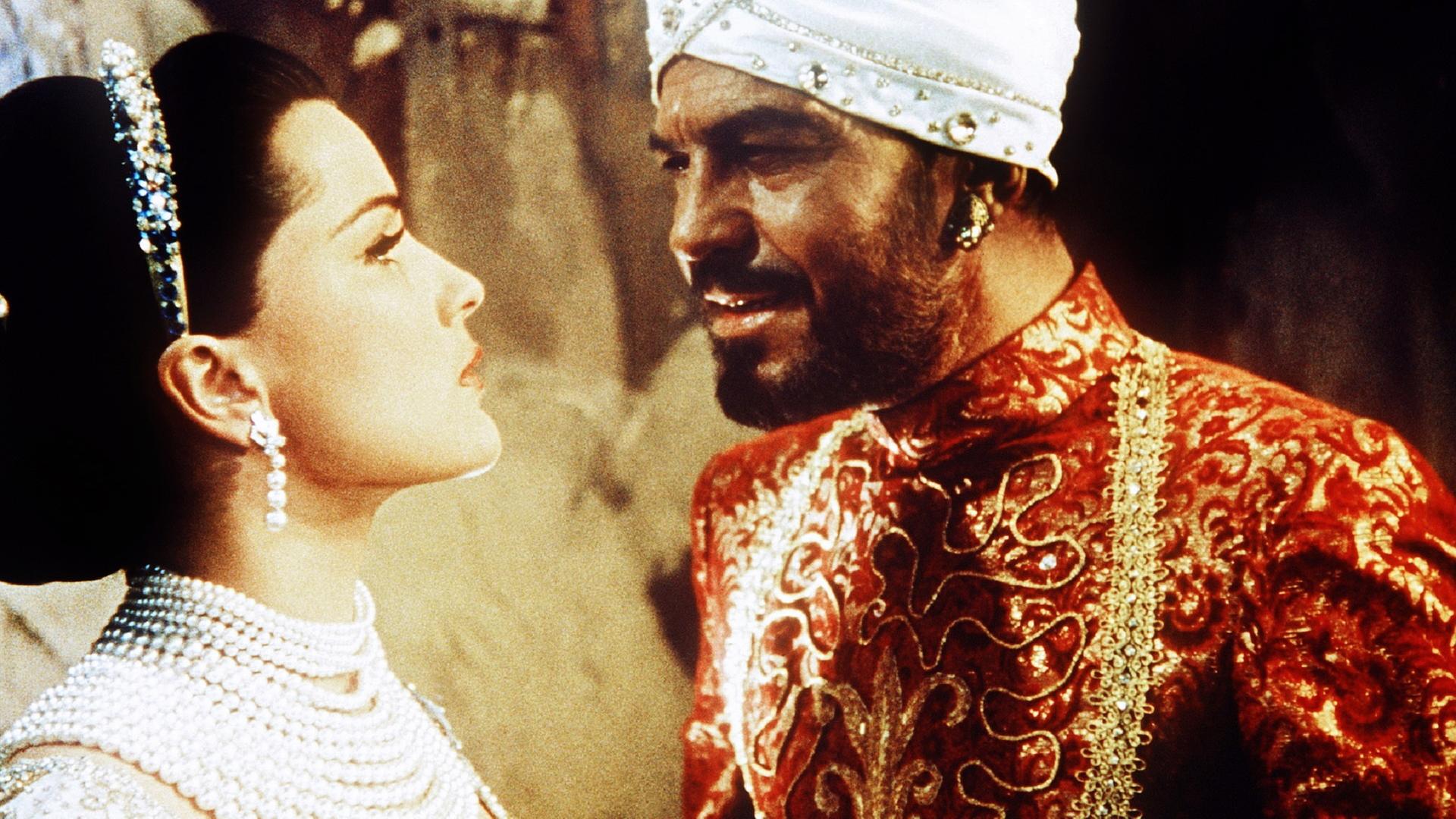 Ein dunkelhäutiger, bärtiger Mann mit weißem Turban nähert sich mit spöttischem Blick von rechts einer festlich hell gekleideten Frau mit halblangem braunem Haar, die mit zahlreichen prunkvollen Perlenketten geschmückt ist und ihn eingeschüchtert anblickt.
