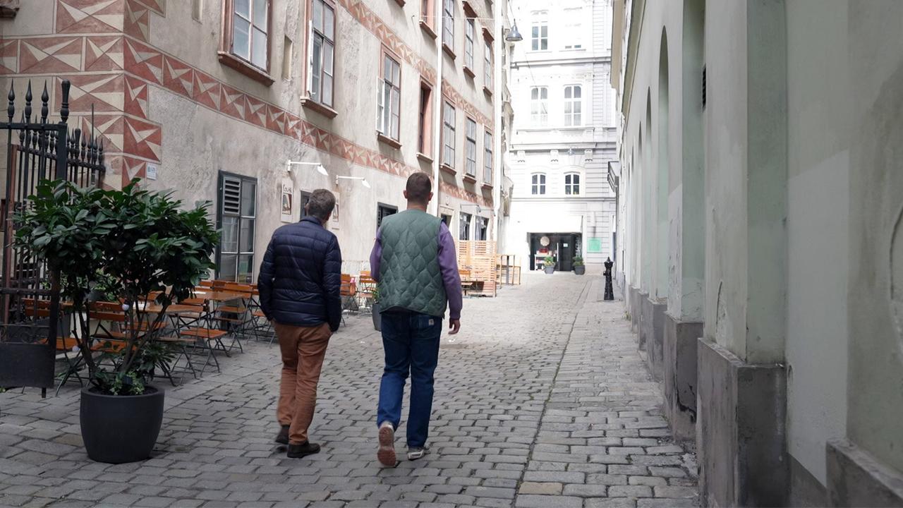 Das Bild zeigt eine enge, gepflasterte Gasse in Wien. Auf der linken Seite befinden sich einige Holztische und Stühle, die offenbar zu einem Café oder Restaurant gehören. Vor den Tischen steht ein großer, runder Topf mit einer Pflanze. An der Wand des Gebäudes sind dekorative Muster in Erdtönen zu sehen.  Zwei Männer, die von hinten zu sehen sind, gehen die Gasse entlang. Der erste Mann trägt eine dunkle Jacke, beige Hosen und hat eine lockige Frisur. Der zweite Mann trägt eine grüne Weste über einem lilafarbenen Pullover und hat kurze, dunkle Haare. Am Ende der Gasse ist ein helles Gebäude mit großen Fenstern sichtbar, das den Eindruck eines weiteren Geschäfts oder eines öffentlichen Gebäudes vermittelt.  Die Gasse hat einen historischen Charakter, umrahmt von älteren Gebäuden, die teilweise wertvolle architektonische Details aufweisen. Die Lichtverhältnisse sind eher gedämpft, was auf eine bewölkte Atmosphäre hindeutet.