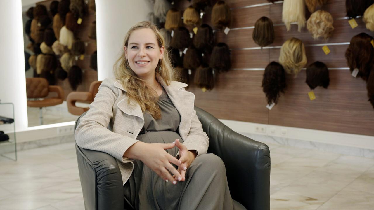 Eine Frau sitzt in einem Sessel vor einer Wand, an der lauter Perücken hängen.