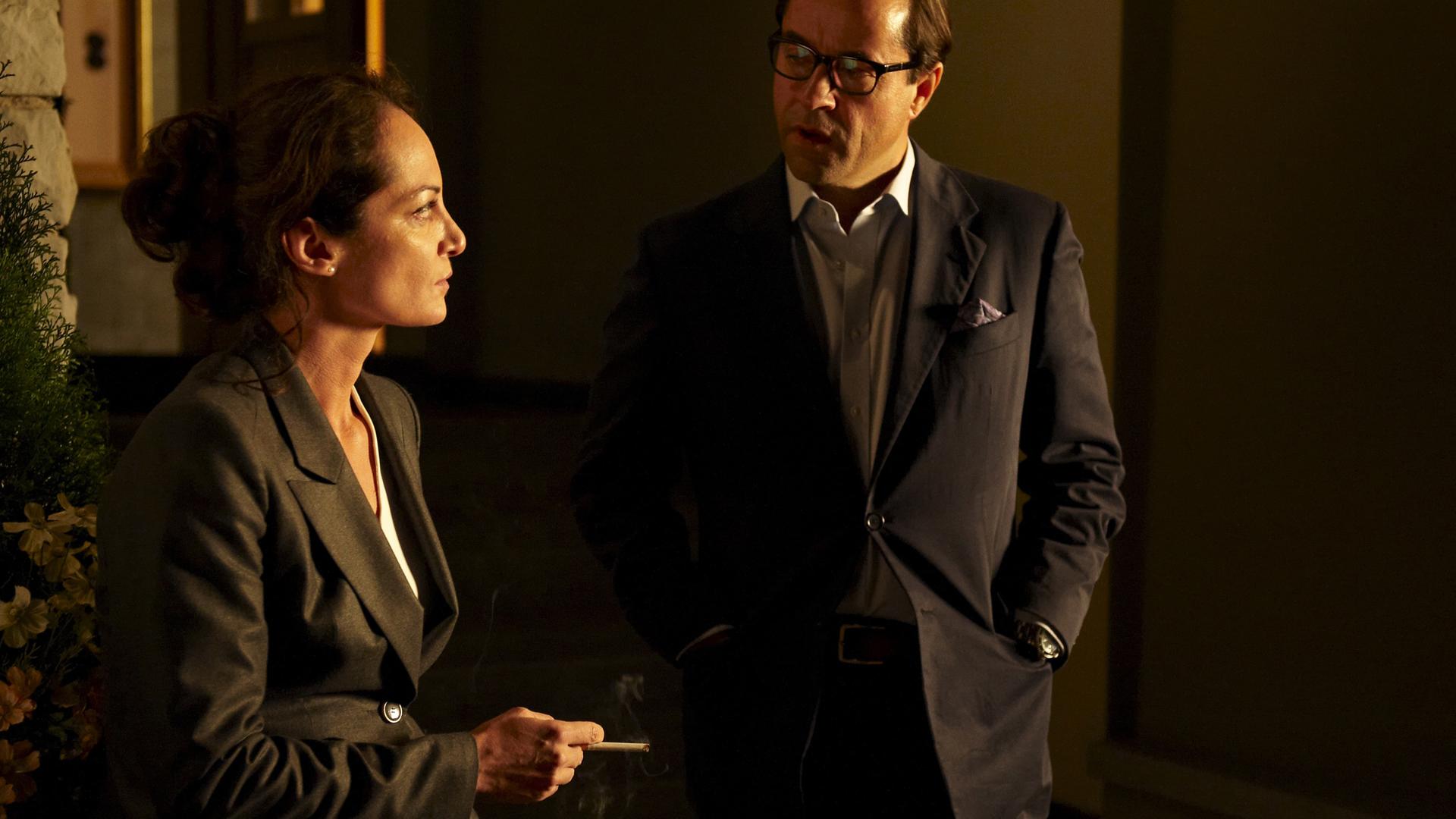 Sigrun (Natalia Wörner) und Vernau (Jan Josef Liefers) stehen nachts vor einem Haus und unterhalten sich. Sigrun hält eine Zigarette in der Hand.