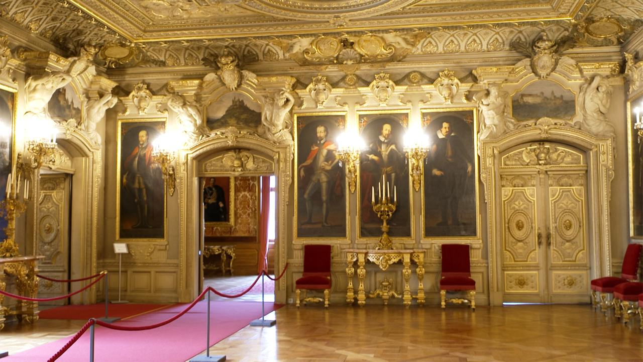 Das Bild zeigt den Innenraum eines prächtigen Schlosses, vermutlich Schloss Sigmaringen. Der Raum ist opulent gestaltet mit reich verzierten Wänden, die sowohl Stuckarbeiten als auch große Gemälde zeigen.   An den Wänden hängen mehrere Portraits, die vermutlich historische Persönlichkeiten darstellen. Der Boden ist aus polierten Holzplanken und hat ein klassisches Fischgrätmuster. In der Mitte des Raums steht ein eleganter Tisch mit einem goldenen Unterbau und Kerzenständern.   Es gibt zwei rote, gepolsterte Stühle, die seitlich des Tisches platziert sind. An der Decke hängen mehrere opulente Leuchten, die den Raum beleuchten. Die Wände sind in warmen, goldenen und beigen Tönen gehalten, was dem Raum ein luxuriöses Ambiente verleiht. Eine große, verzierte Tür an einer Wand führt möglicherweise zu einem weiteren Raum.   Im Hintergrund sind Teile des Raumes sichtbar, die für weiteres Interesse sorgen, darunter eine offene Tür, die zu einem anderen Bereich des Schlosses führt. Der gesamte Raum vermittelt den Eindruck von historischem Reichtum und Eleganz.