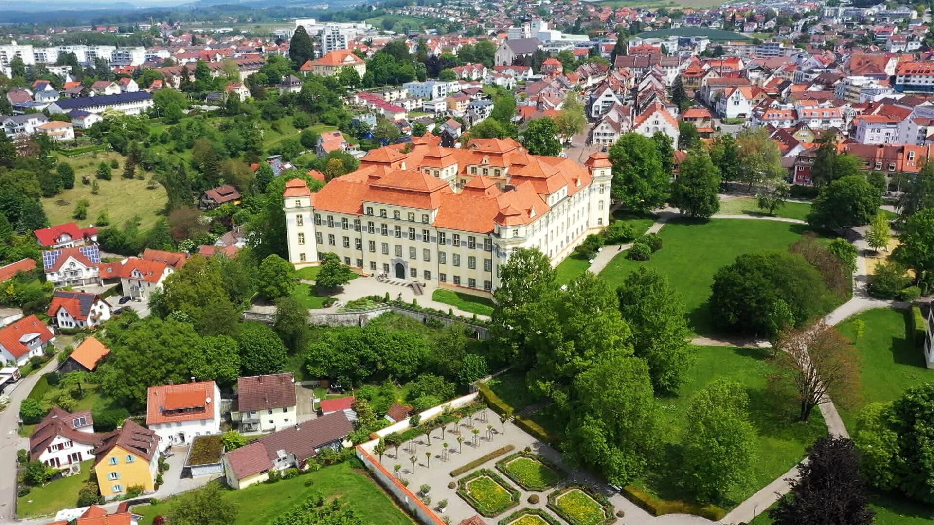 Das Bild zeigt das Schloss Tettnang in einer anschaulichen Perspektive. Das Schloss hat eine helle Fassade und ein markantes, rot gedecktes Dach mit mehreren Erkern und Türmen. Vor dem Schloss erstreckt sich ein gepflegter Garten mit symmetrischen Blumenbeeten, während um das Schloss herum Rasenflächen und Bäume zu sehen sind.  Im Hintergrund sind Teile der Stadt Tettnang zu erkennen, mit vielen kleinen Gebäuden und Wohnhäusern, die ebenfalls rote Dächer besitzen. Diese Gebäude sind in unterschiedlicher Höhe angeordnet und einige sind von Bäumen umgeben. Von dieser Perspektive aus bietet sich auch ein Blick auf die hügelige Landschaft und den klaren Himmel im Hintergrund.  Das Bild vermittelt ein Gefühl von historischer Pracht und zeigt die Verbindung zwischen der Natur und der Architektur des Barockzeitalters.