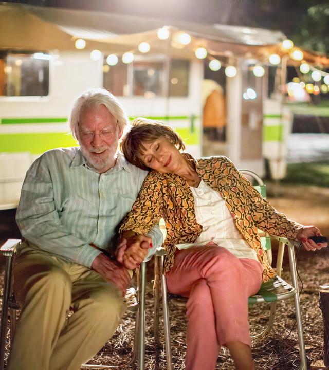 Ein älterer Mann und eine ältere Frau sitzen bei Dunkelheit auf dem Camping-Platz dicht zusammen, halten sich gegenseitig die Hand und schauen sich Dias an. Sie wirken glücklich und zufrieden. Im Hintergrund ist ein mit einer Lichterkette beleuchtetes Wohnmobil zu sehen.