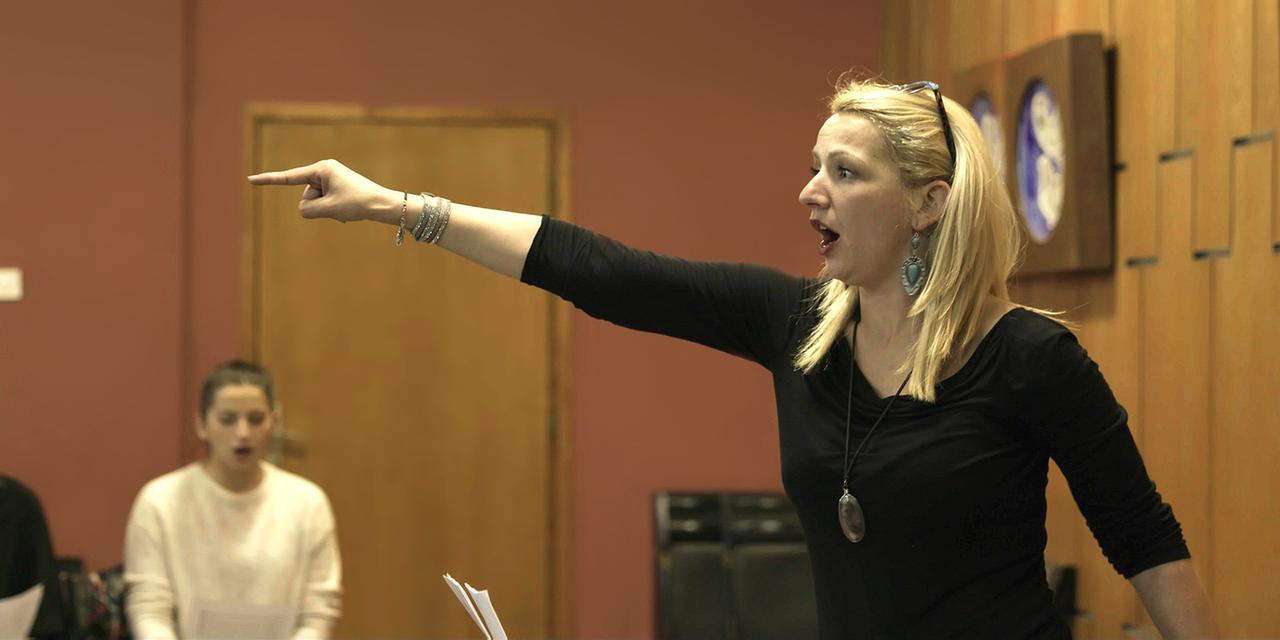 Eine Frau mit langen blonden Haaren zeigt bei einer Chorprobe in einer lebhaften Geste mit ausgestrecktem Arm und Finger auf eine Person außerhalb des Bildes.