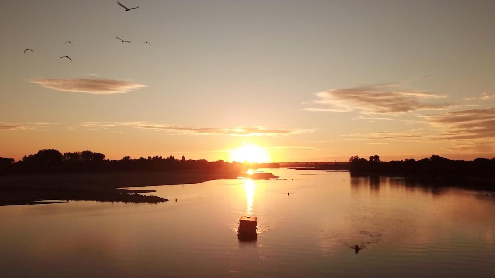 Panoramaaufnahme der Loire mit ihrem breiten Flussbett: Die untergehende Sonne taucht die Landschaft in rötliches Licht. Am Himmel hängen einige Wolken. In der Mitte des Flusses fährt ein Boot.
