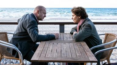 Zwei Personen sitzen sich an einem Holztisch auf einer Strandterrasse gegenüber, im Hintergrund Meer und Sandstrand – beide blicken sich ernst an.