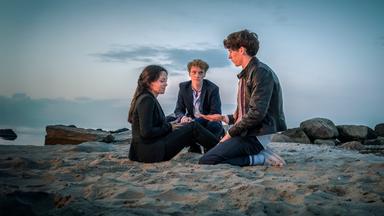 Drei junge Menschen sitzen in feiner Kleidung am Strand im Abendlicht – sie wirken nachdenklich, das Meer und Felsen im Hintergrund.