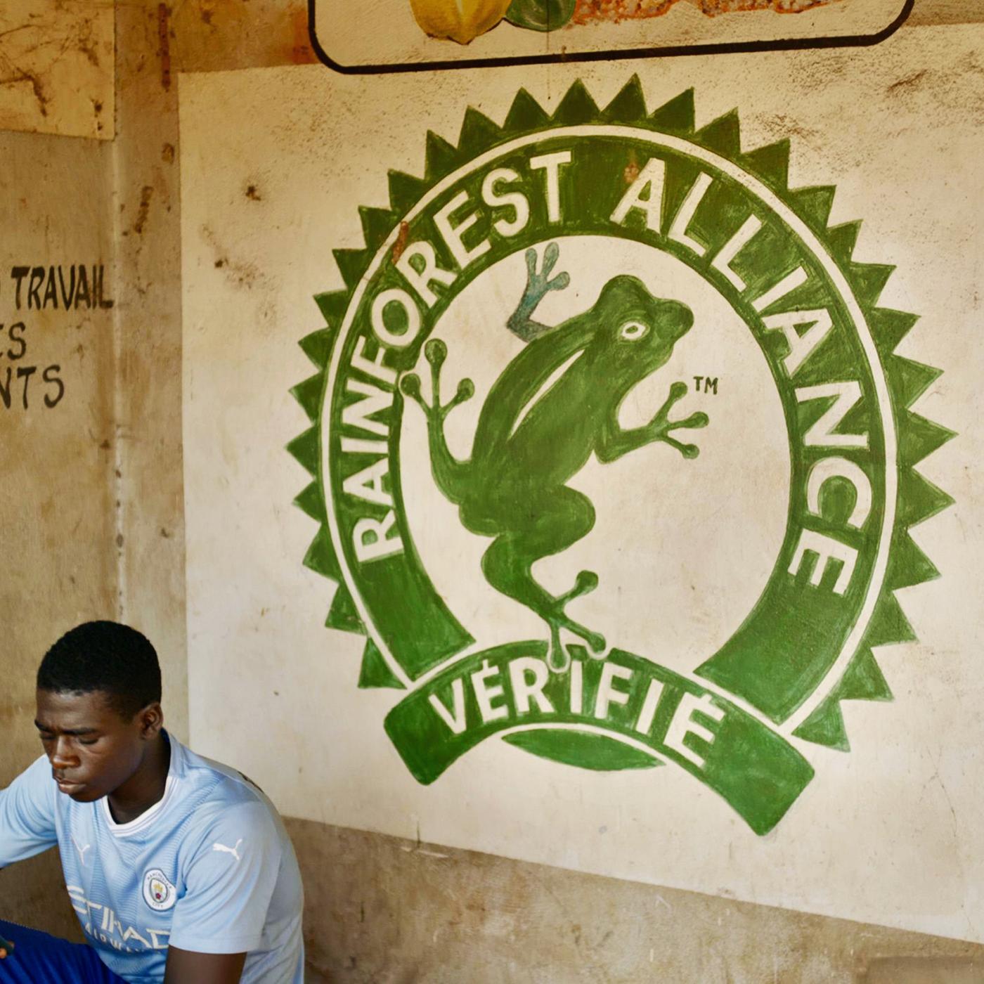 Ein junger Mann sitzt in einem Raum und schaut auf sein Mobiltelefon. Hinter ihm sieht man eine Wand mit einem Logo.