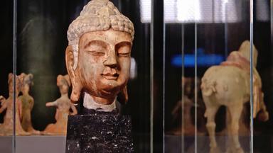 Ein Buddha-Kopf in einer Vitrine. Im Hintergrund weitere Kunstwerke.