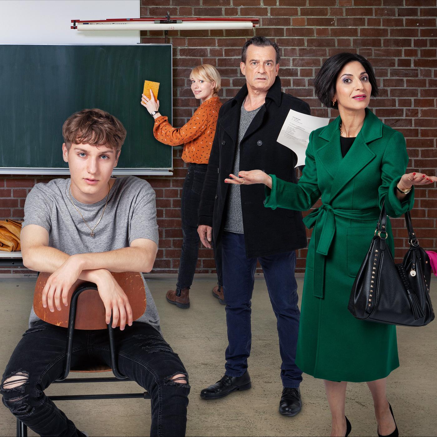 Eine Schultafel, ein Tisch, sieben Erwachsene und ein Jugendlicher im Vordergrund sind zu sehen.