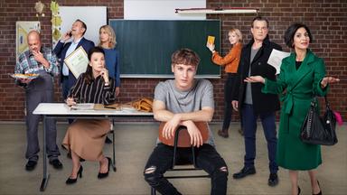 Eine Schultafel, ein Tisch, sieben Erwachsene und ein Jugendlicher im Vordergrund sind zu sehen.