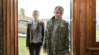 Das Bild zeigt zwei Personen, die in einem Türrahmen stehen. Auf der linken Seite ist eine junge Frau zu sehen. Sie trägt eine schwarze Lederjacke über einem grauen Rollkragenpullover und hat lange, glatte Haare. Ihre Miene wirkt ernst und konzentriert. Neben ihr steht ein Mann, der älter aussieht. Er trägt eine olivfarbene Jacke über einem grauen T-Shirt und schaut ebenfalls ernst.   Durch den Türrahmen ist eine grüne Wiese und im Hintergrund sind Bäume und Berge sichtbar. Hinter den Personen ist ein Auto zu erkennen, das teilweise im Bild ist. Die Stimmung wirkt angespannt und nachdenklich.