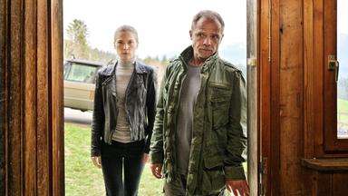 Das Bild zeigt zwei Personen, die in einem Türrahmen stehen. Auf der linken Seite ist eine junge Frau zu sehen. Sie trägt eine schwarze Lederjacke über einem grauen Rollkragenpullover und hat lange, glatte Haare. Ihre Miene wirkt ernst und konzentriert. Neben ihr steht ein Mann, der älter aussieht. Er trägt eine olivfarbene Jacke über einem grauen T-Shirt und schaut ebenfalls ernst.   Durch den Türrahmen ist eine grüne Wiese und im Hintergrund sind Bäume und Berge sichtbar. Hinter den Personen ist ein Auto zu erkennen, das teilweise im Bild ist. Die Stimmung wirkt angespannt und nachdenklich.