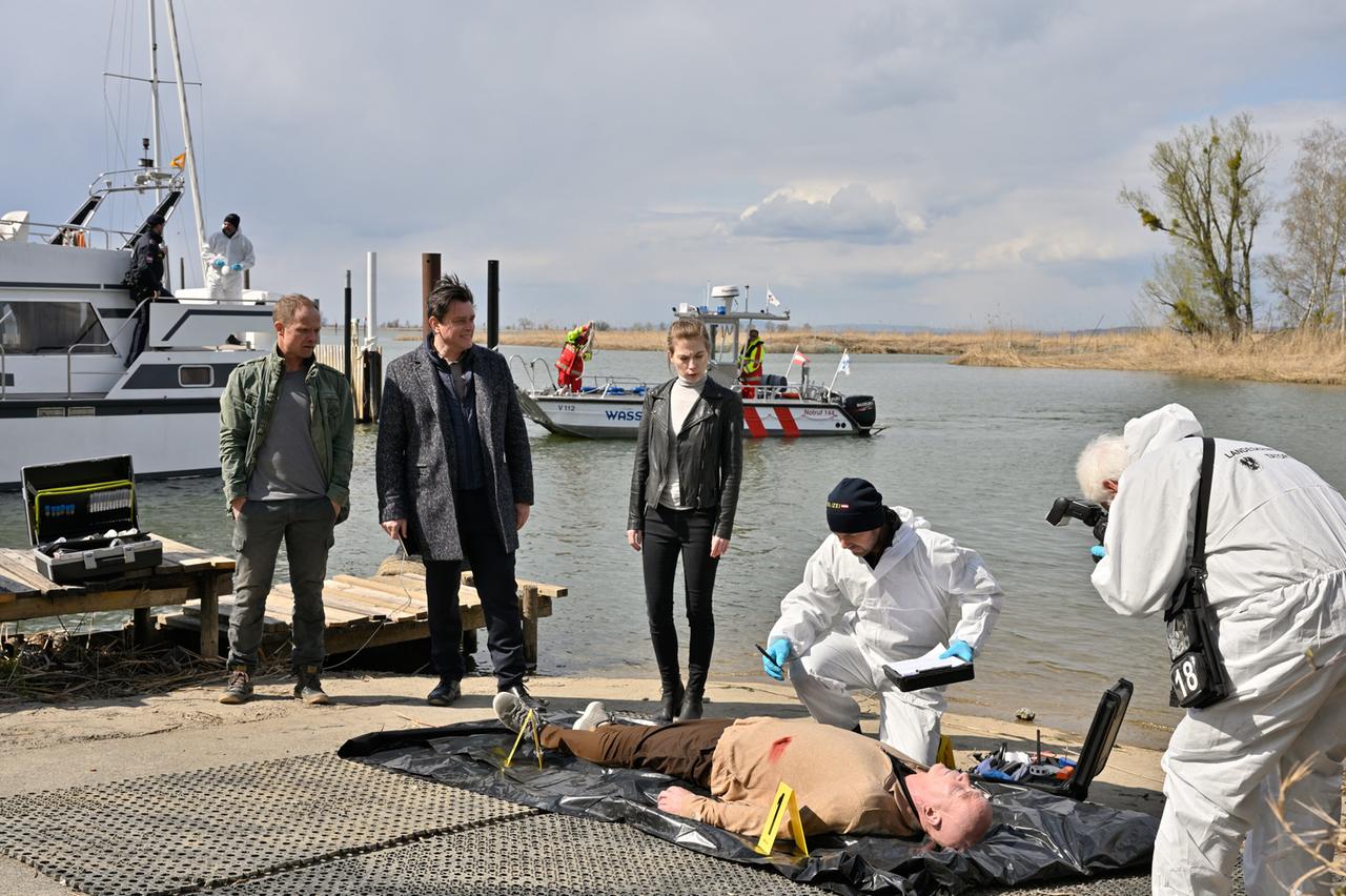 Das Bild zeigt eine Szene am Ufer eines Gewässers, wahrscheinlich einem See. Im Vordergrund liegt ein männlicher Körper auf einem schwarzen Folienuntergrund. Der Mann trägt einen beigefarbenen Pullover und hat blutige Wunden auf der Brust. Rund um ihn sind gelbe Markierungen angebracht, die in einem Kriminalitätskontext üblich sind.  Links im Bild stehen zwei Männer: Der erste trägt eine grüne Jacke und hellgraue Hose, der zweite hat einen dunklen Mantel an. Beide blicken auf den Körper. Rechts steht eine Frau in schwarzer Lederjacke und heller Bluse, die ebenfalls auf den Körper schaut.  Im Hintergrund ist ein Polizei- bzw. Rettungsboot zu sehen, auf dem mehrere Personen in Schutzkleidung arbeiten. Zusätzlich sind hinter dem Boot einige Holzpfähle und Schilf zu erkennen, was auf das Ufer des Sees hinweist. Der Himmel ist bewölkt und es scheint, als könnte es bald regnen.   Insgesamt vermittelt das Bild eine spannende und ernste Atmosphäre, die auf eine kriminalistische Untersuchung hinweist.