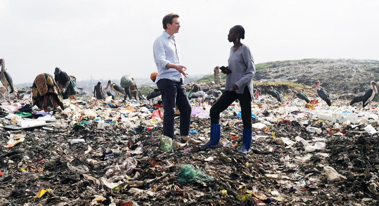 Ein Mann und eine Frau stehen in einer Mülldeponie zwischen Menschen und Tieren, die im Müll suchen.