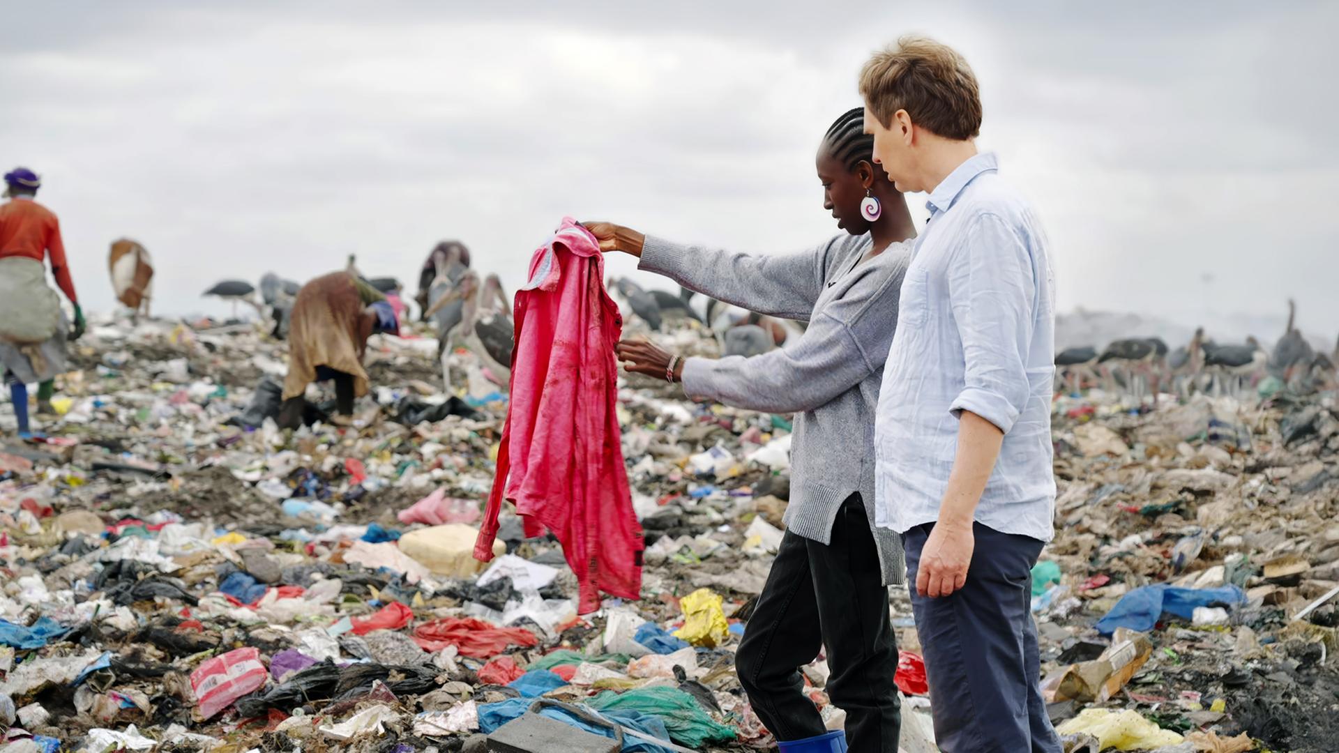 Ein Mann und eine Frau stehen in einer Mülldeponie zwischen Menschen und Tieren, die im Müll suchen.
