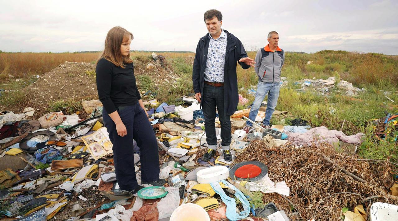 Zwei Männer und eine Frau stehen zwischen Müll auf einer Wiese