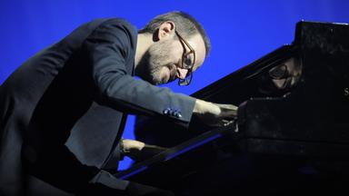 Ein Mann mit Brille und Bart beugt sich über ein Klavier und spielt.