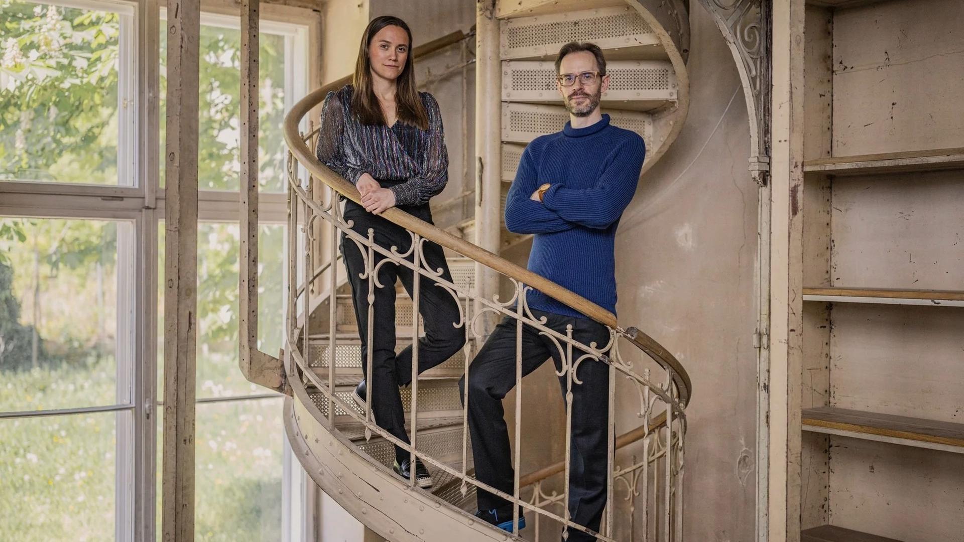 Zwei Musiker stehen in einem Innenraum mit Instrumenten; das Duo ist nebeneinander positioniert, die Umgebung wirkt wie ein Studio oder Proberaum mit sichtbaren Details im Hintergrund.