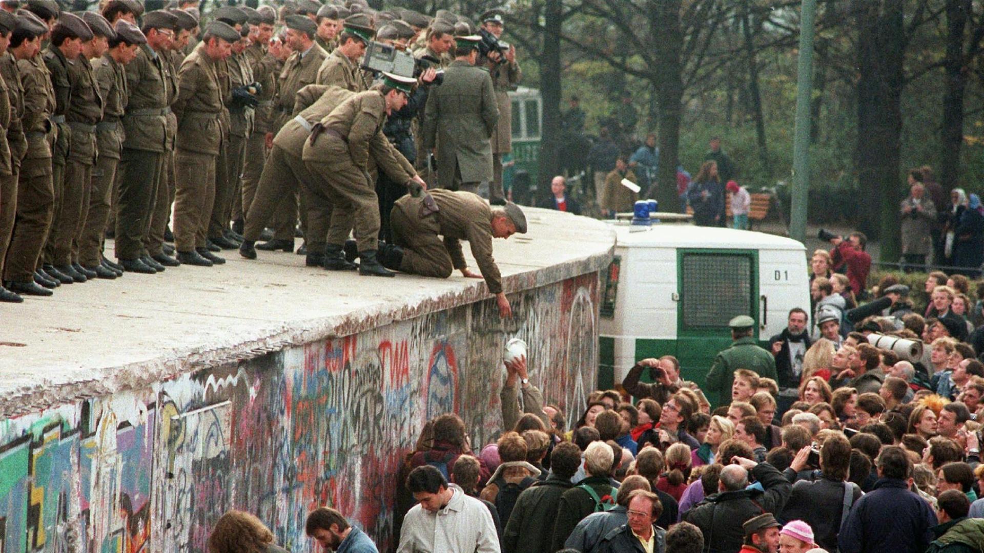 Mehrere DDR-Grenzposten und einer von ihnen bekommt von einem Demonstranten eune Kanne Kaffe auf die Mauer vor dem Brandenburger Tor gereicht.